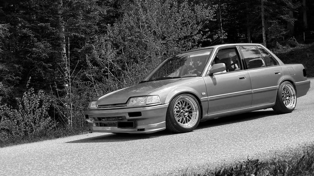 Hochwertiges Schwarz-Weiß-Foto eines modifizierten, tiefergelegten klassischen Honda Civic EF Sedan auf einer asphaltierten Waldstraße. Das Fahrzeug zeigt polierte Mesh-Felgen, eine Frontlippe und ist vor einem dichten Waldhintergrund positioniert
