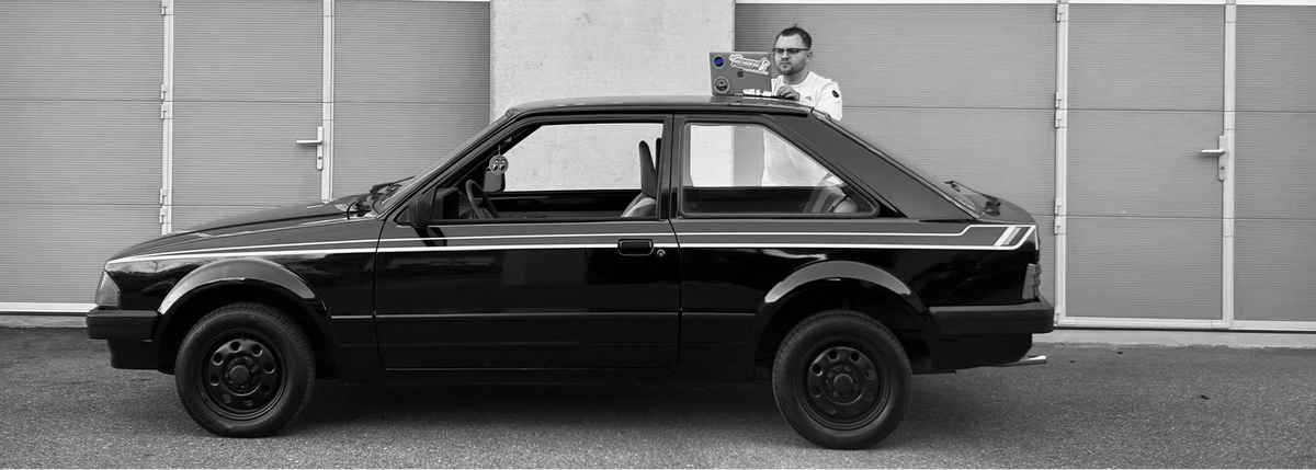 Schwarz-Weiß-Panoramafoto für die Über Uns Seite von MediaMechs.ch. Ein Teammitglied mit Brille steht hinter einem gepflegten, klassischen schwarzen Ford Escort (Oldtimer Hatchback) mit weißen Seitenstreifen. Ein offener Laptop ruht auf dem Dach des Autos, während die Person in die Kamera blickt. Der Hintergrund zeigt eine Reihe von industriellen Garagentoren. Das Bild symbolisiert die Verbindung von moderner Technik und klassischem Automobil-Handwerk.