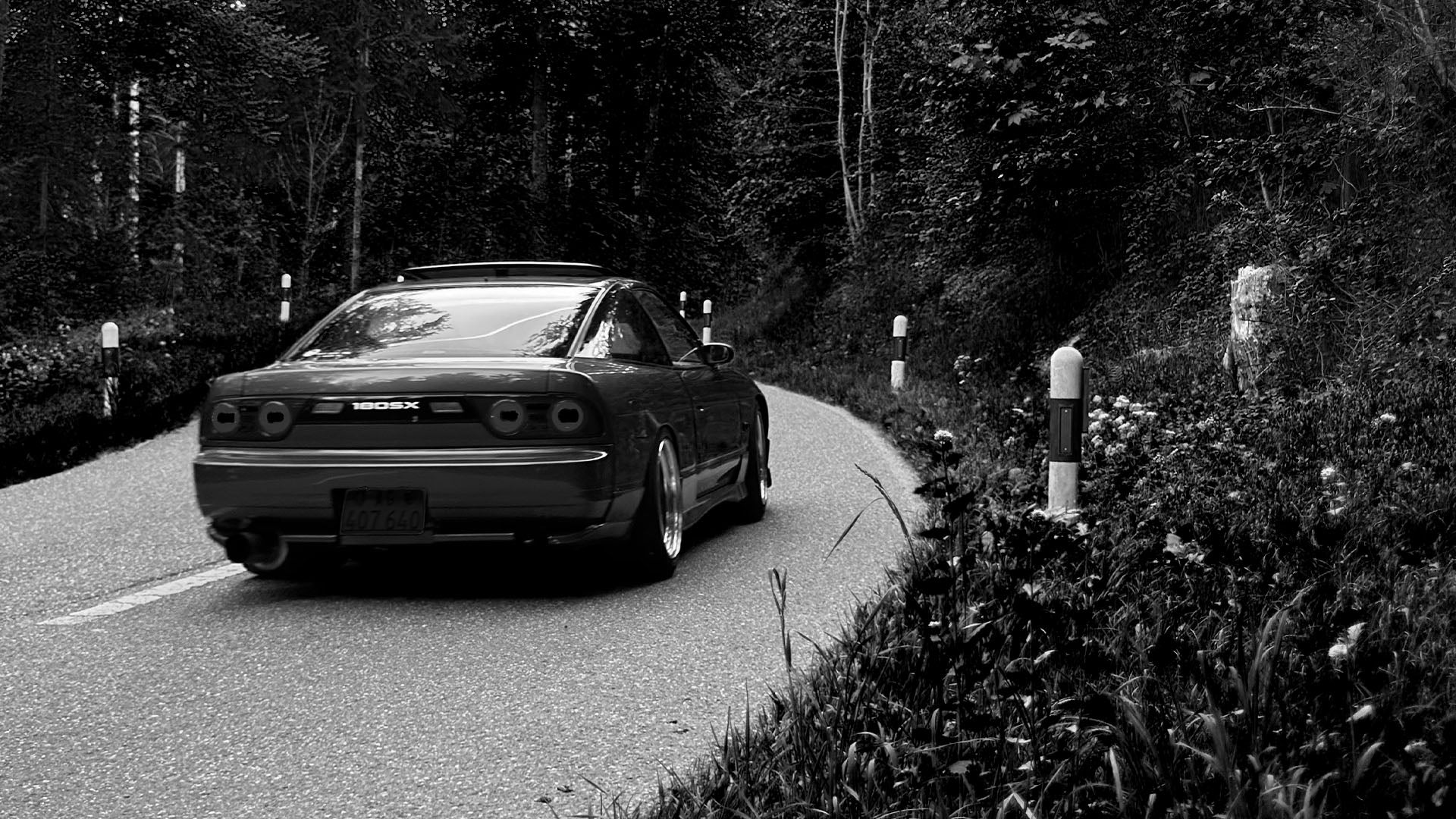 Stimmungsvolle Schwarz-Weiß-Aufnahme, die die Heckansicht eines modifizierten Nissan 180SX auf einer kurvenreichen Bergstraße zeigt. Die JDM-Kultikone fährt durch einen dichten Wald. Details wie der lesbare 180SX-Schriftzug, Aftermarket-Rückleuchten, abgesenktes Fahrwerk und große Chromfelgen sind sichtbar, flankiert von Begrenzungspfosten und wilden Gräsern.