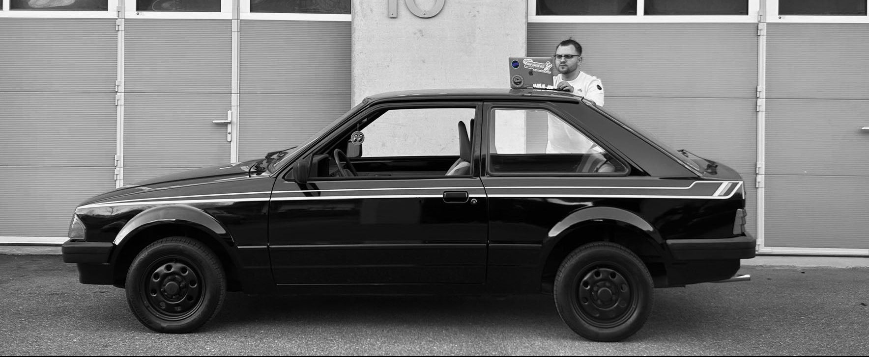 Digitales Fahrzeug-Marketing direkt am Objekt: Media Mechs Experte arbeitet am Laptop auf dem Dach eines schwarzen Ford Escort MK3 vor einer Werkstatt-Halle